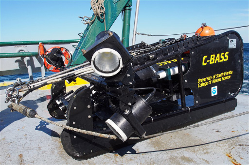 A towed camera rests on a boat with rigging, flotation devices, ocean, and blue sky visible in the background. The towed camera is a large piece of equipment.